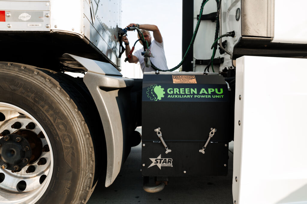 Green APU Star Edition diesel APU mounted on the frame rail of a white semi truck while the driver hooks up trailer lines